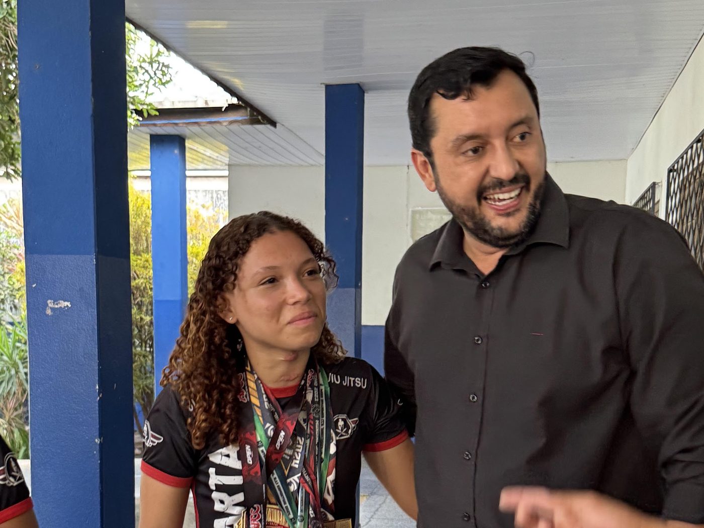 Allan Kardec sorri ao lado de jovem atleta de jiu-jitsu com várias medalhas no peito, em visita a escola