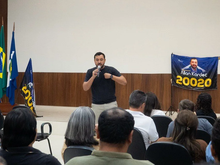 Professor Allan Kardec fala ao microfone em auditório, com bandeira 20020 ao fundo