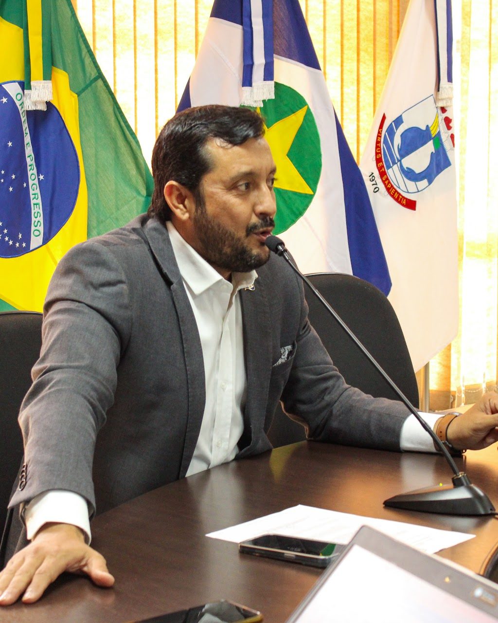 Professor Allan Kardec fala ao microfone em mesa oficial, com as bandeiras do Brasil e de Mato Grosso ao fundo